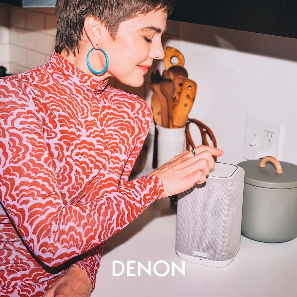 Denon Home wireless speaker on a kitchen counter, being set up by a young woman in a bold red patterned turtleneck and blue hoop earrings; warm domestic lighting, mid-century ceramic canister and wooden utensils nearby.